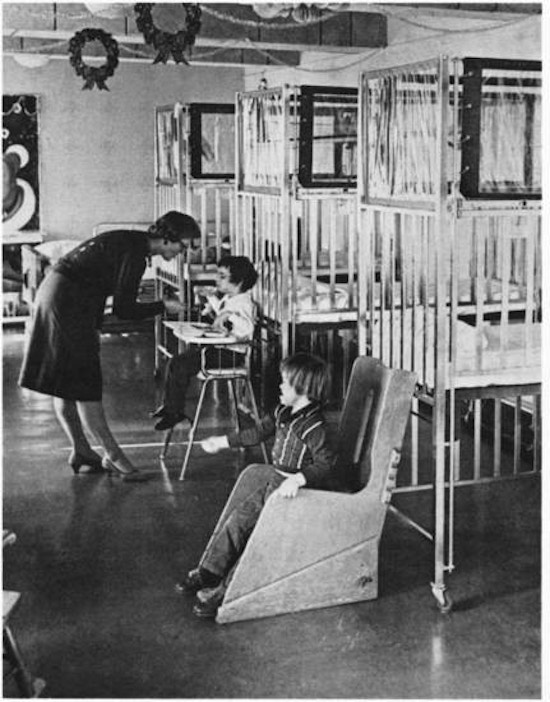 A woman feeding a child in a high chair. The decorated room has cribs.