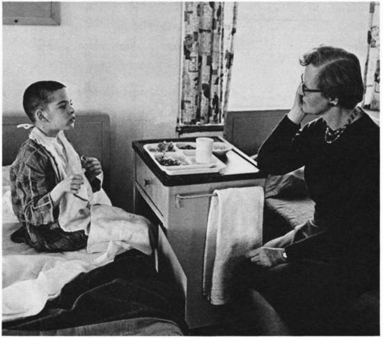 A boy sits on a bed with a tray of food and speaks with a woman.
